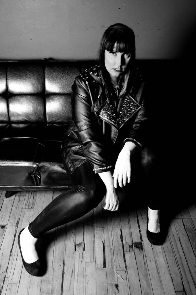 A woman sitting on a leather couch, dressed in a black leather jacket with studs, black leggings, and black flats. The photo is in black and white, showcasing a serious expression.